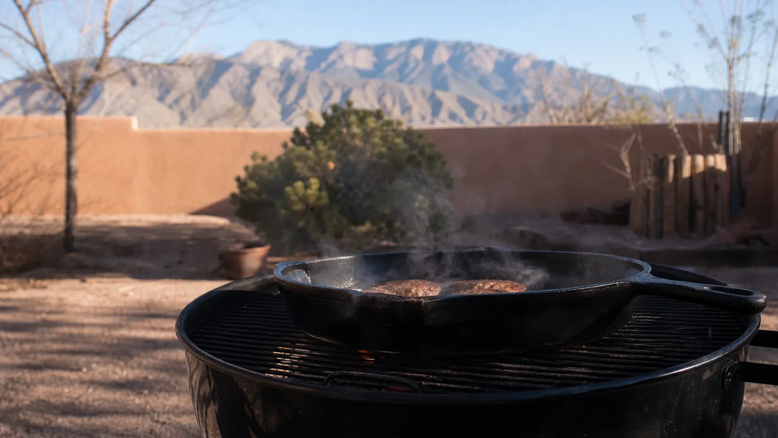 A cast iron griddle sitting over a charcoal kettle grill on a New Mexico patio in early morning light, with the Sandia mountains in the background