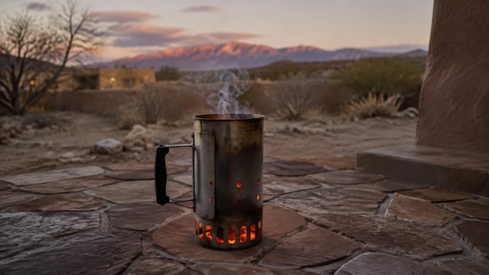 A metal charcoal chimney starter glowing orange at the base with coals lit inside, sitting on a stone patio at dusk