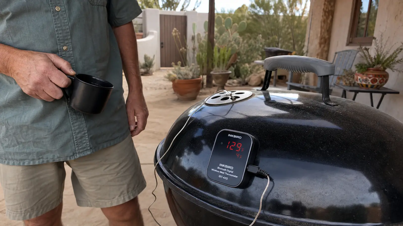 A small digital thermometer base station magnetically mounted to the curved hood of a kettle grill with a probe wire threading through the top vent