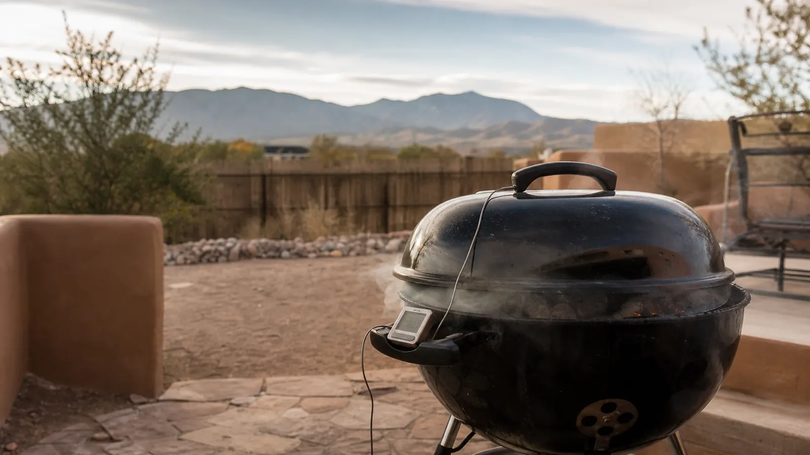 A kettle grill venting smoke at sunrise on a high desert patio with a small wireless thermometer base station clipped to the handle