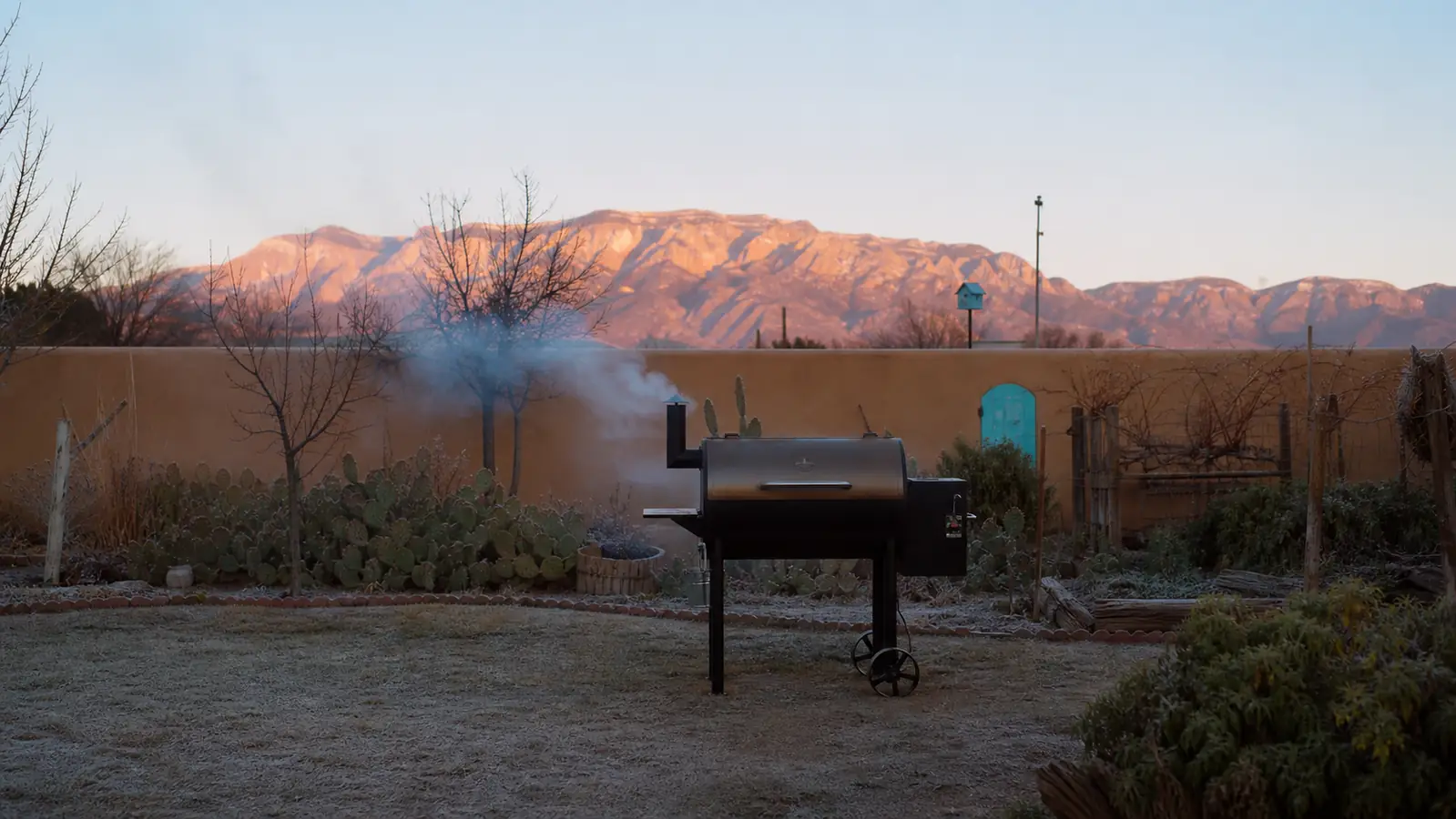A pellet grill smoking in a backyard at dawn with the Sandia Mountains in the background
