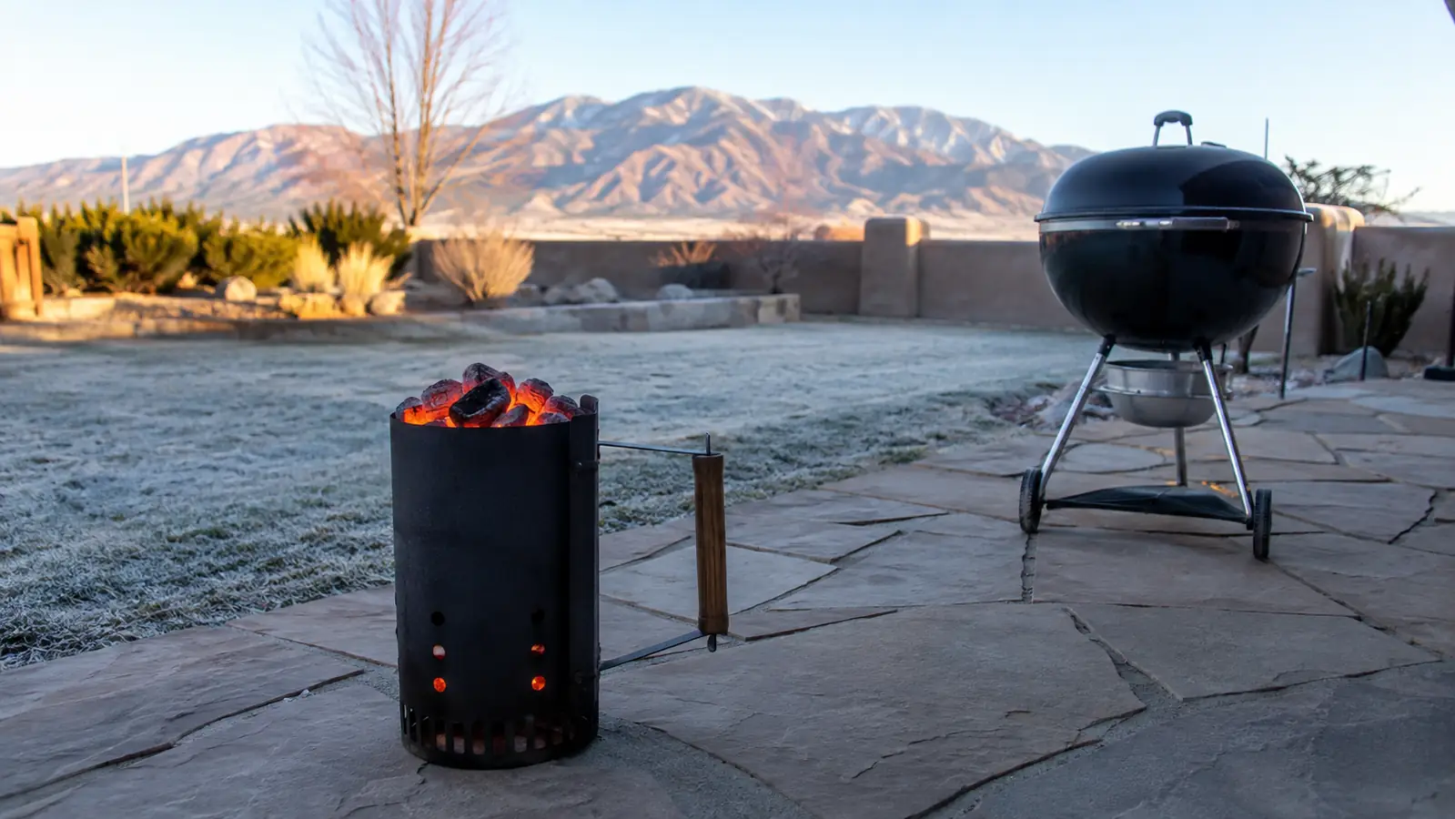 A chimney starter full of glowing orange charcoal sitting on a stone patio in early morning light with a kettle grill nearby