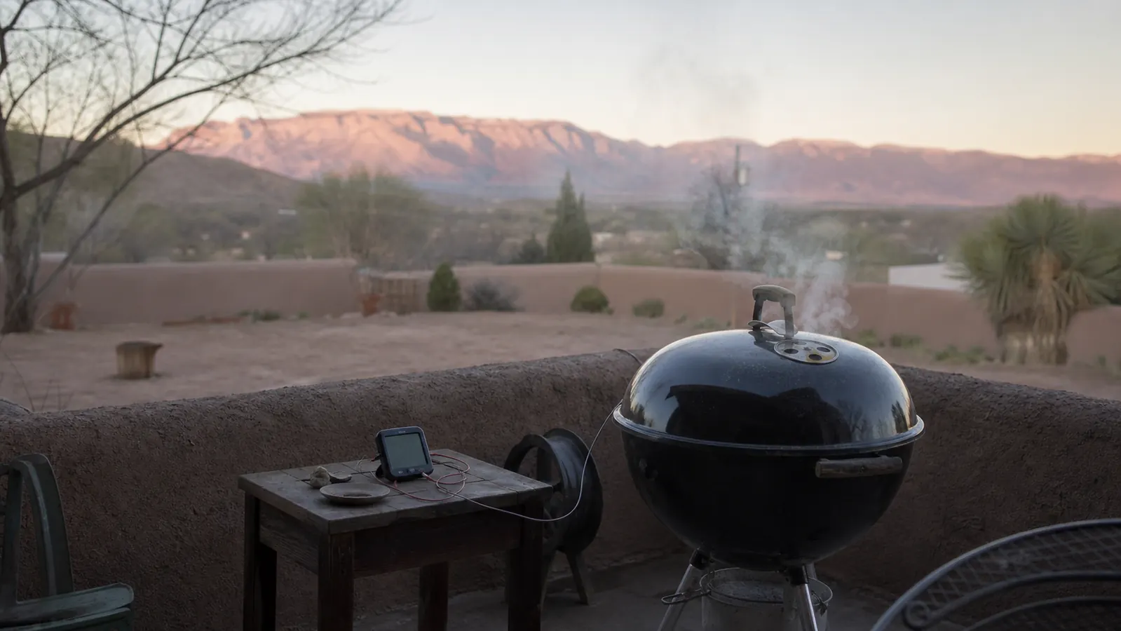 A kettle grill at dawn on a patio with a small wireless thermometer base station sitting on the side table next to it