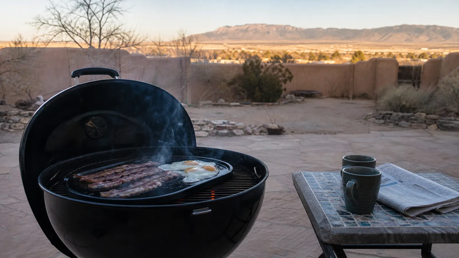 A cast iron griddle steaming on top of a kettle grill on a quiet patio early on a Sunday morning