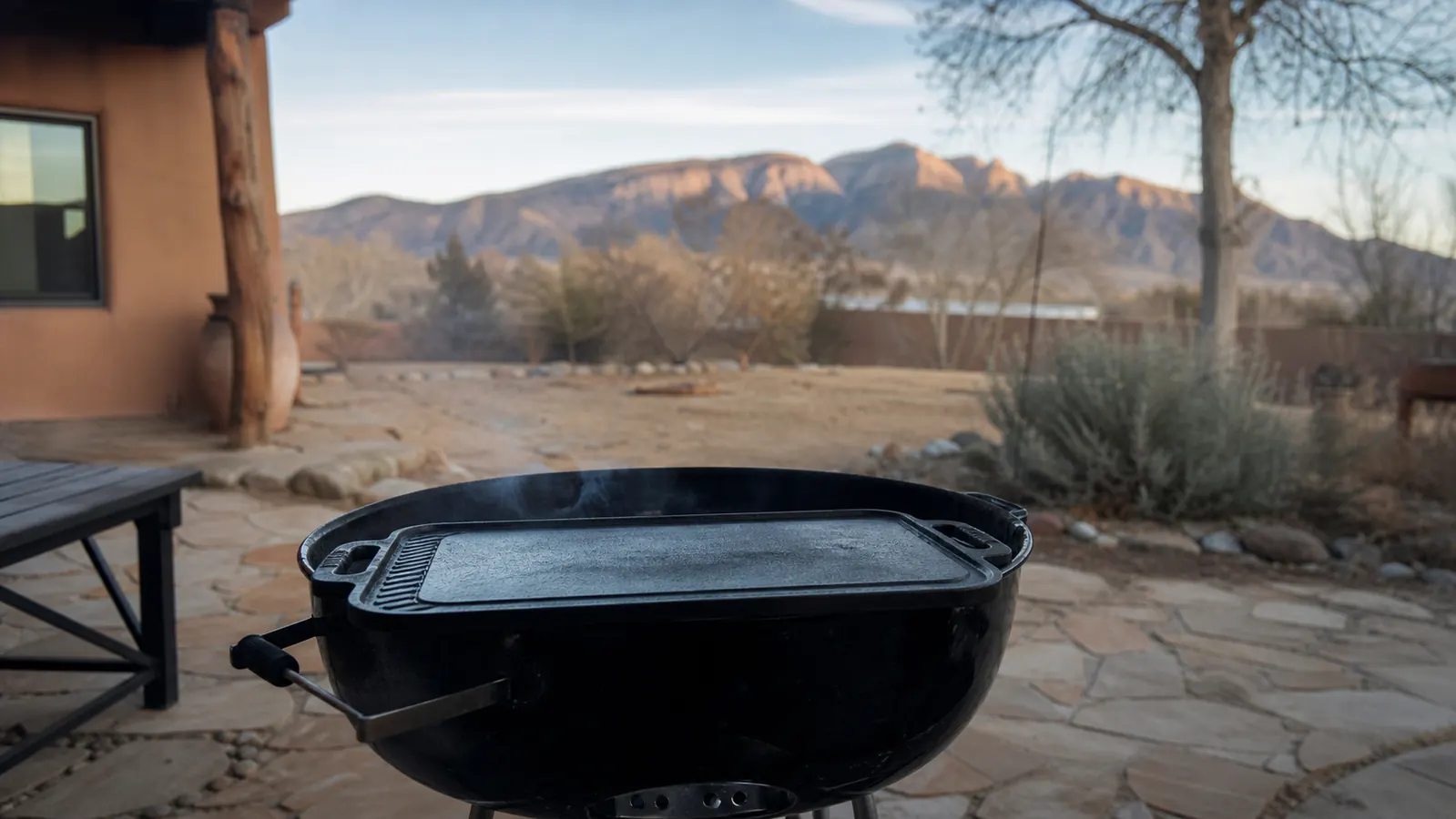 A cast iron griddle sitting on top of the open grates of a kettle grill on a stone patio in early morning light