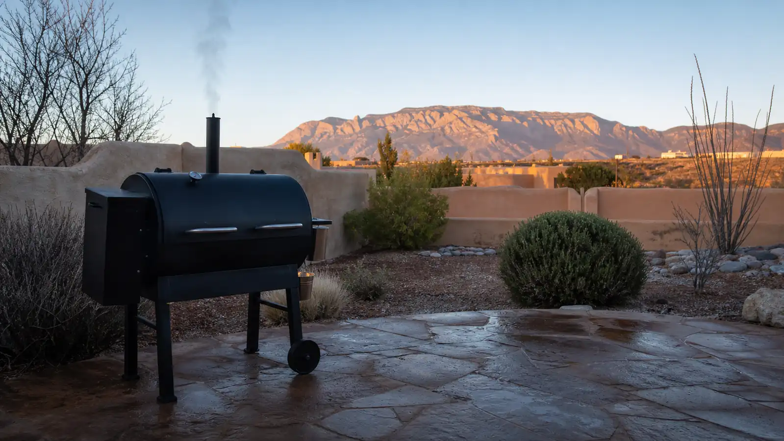 A pellet smoker on a stone patio at dawn with smoke curling from the stack and mountains in the distance