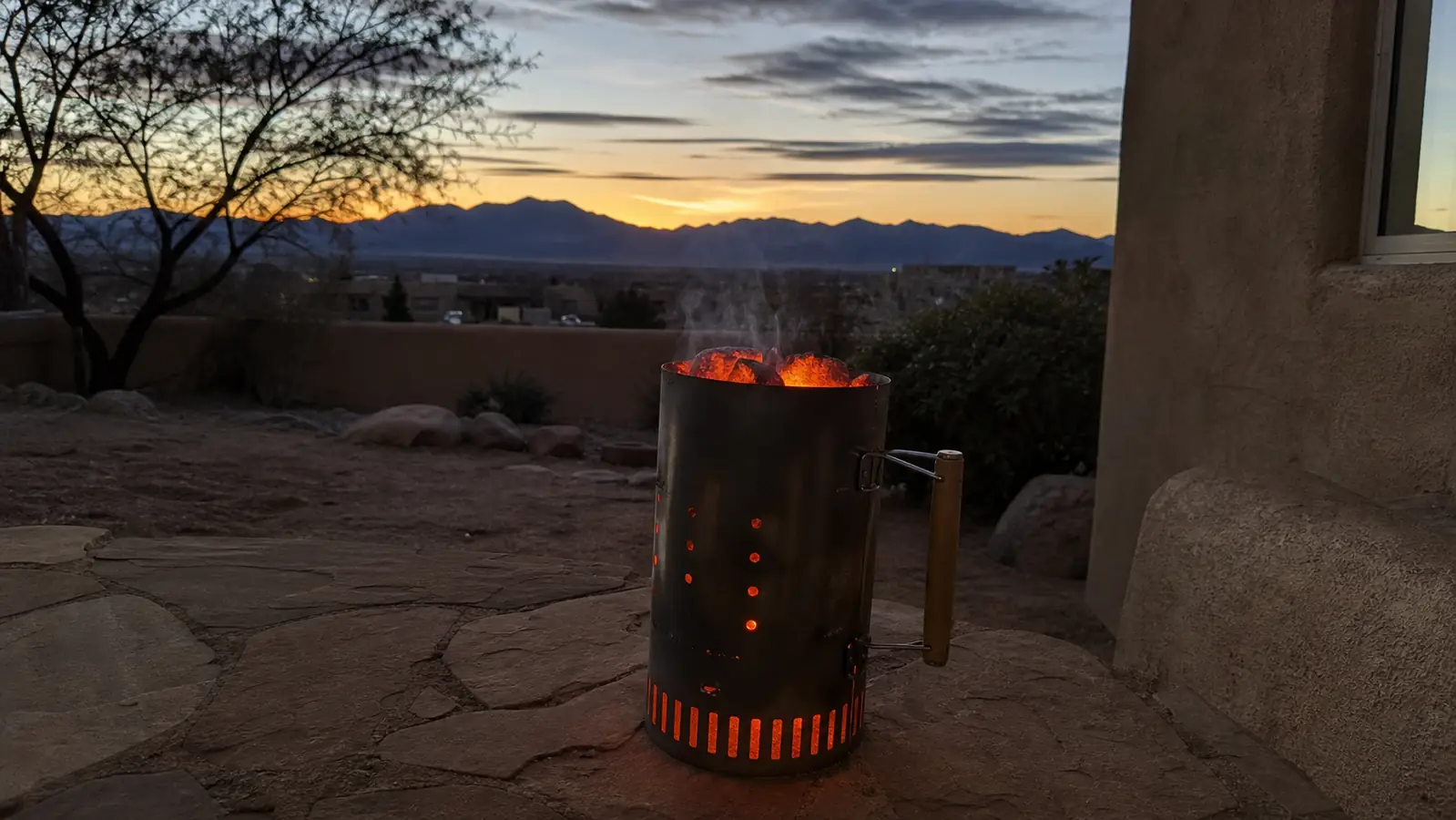 A metal chimney starter full of glowing charcoal sitting on a stone patio at dusk with the New Mexico high desert in the background