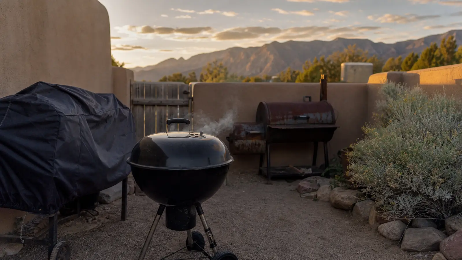 A black kettle grill on a small Albuquerque patio crowded next to a covered gas grill and an old offset smoker at dusk