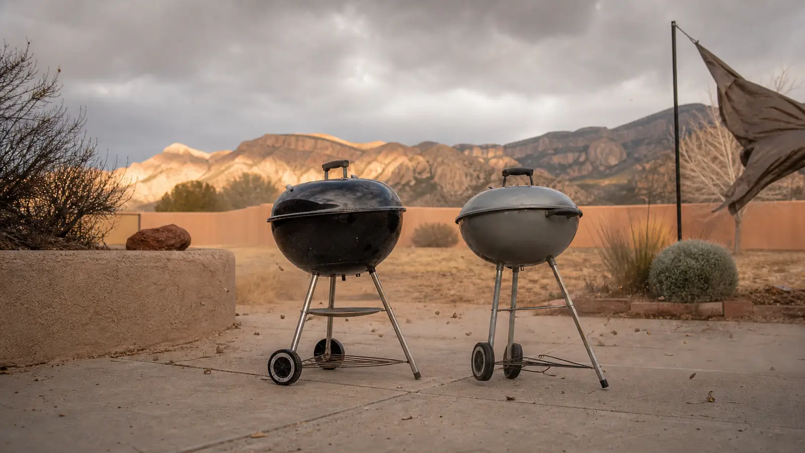 Two charcoal kettle grills side by side in a driveway on a windy overcast afternoon, one heavier black kettle and one lighter thin-steel kettle