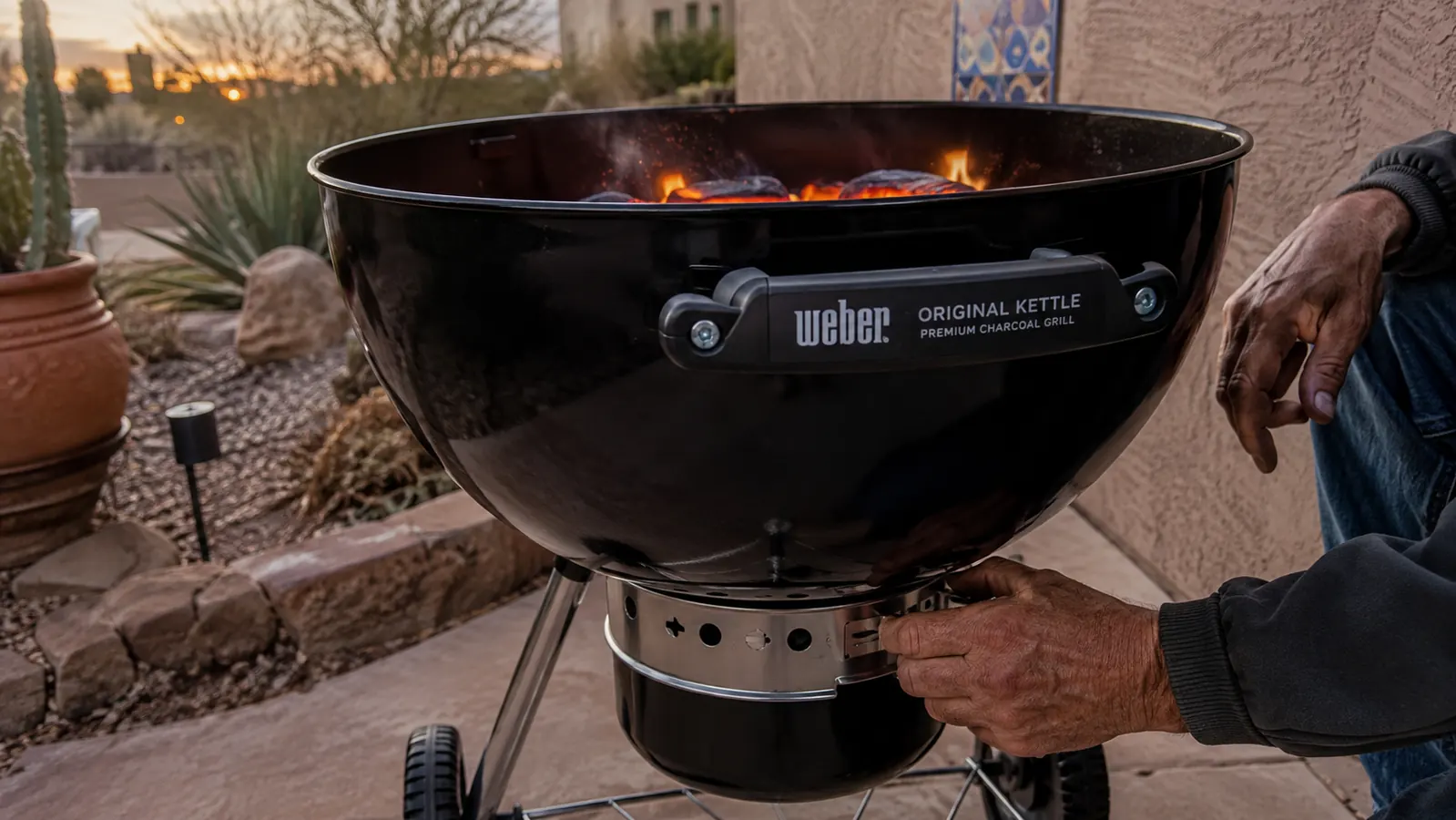 A man's hands adjusting the bottom vent slider on a black kettle grill while coals glow orange inside