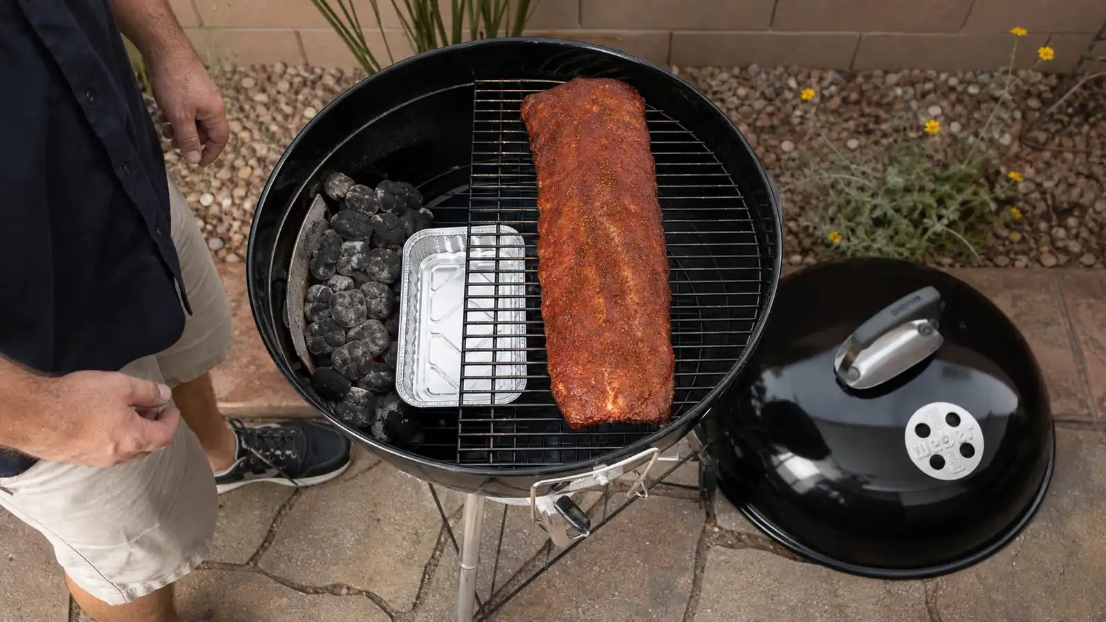 A rack of pork ribs and a foil pan of water arranged on one side of an open kettle grill for indirect cooking