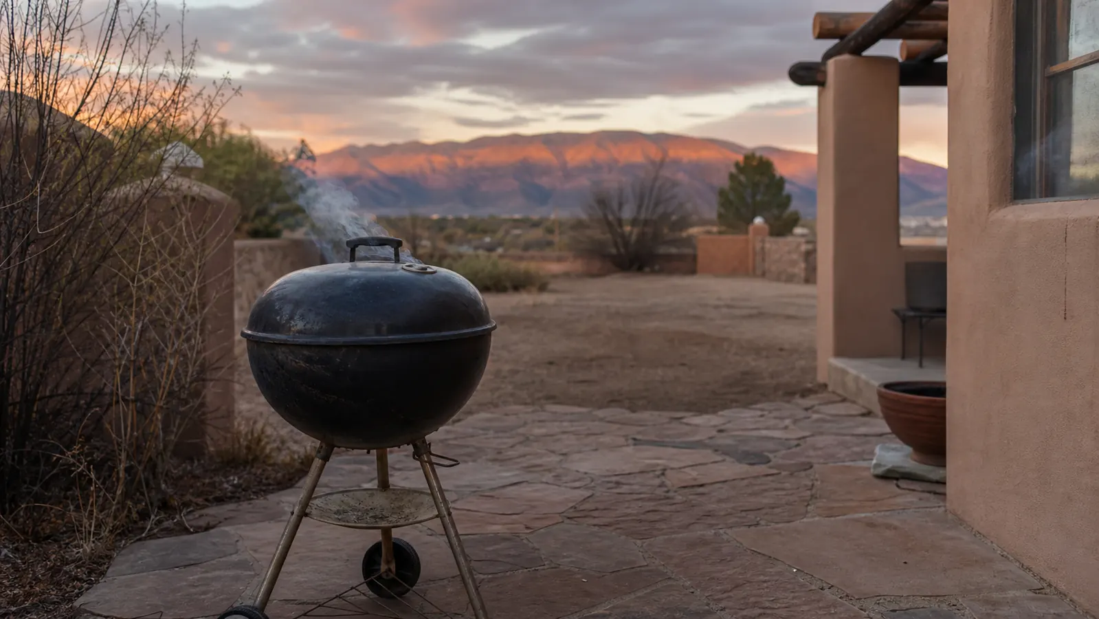 A black Weber kettle grill on a stone patio at golden hour with the Sandia Mountains in the background