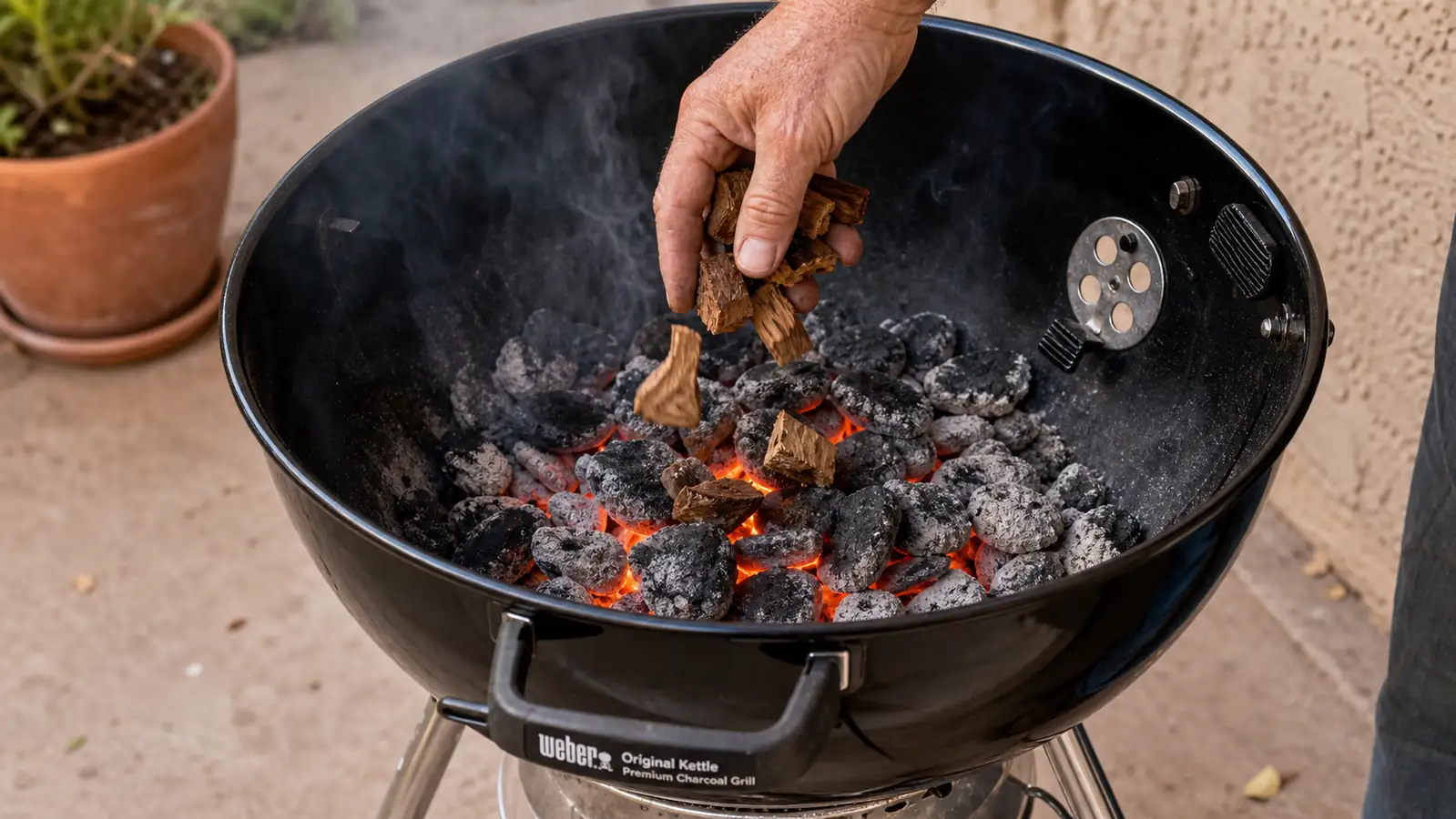 A hand sprinkling mesquite wood chunks onto glowing charcoal inside an open kettle grill